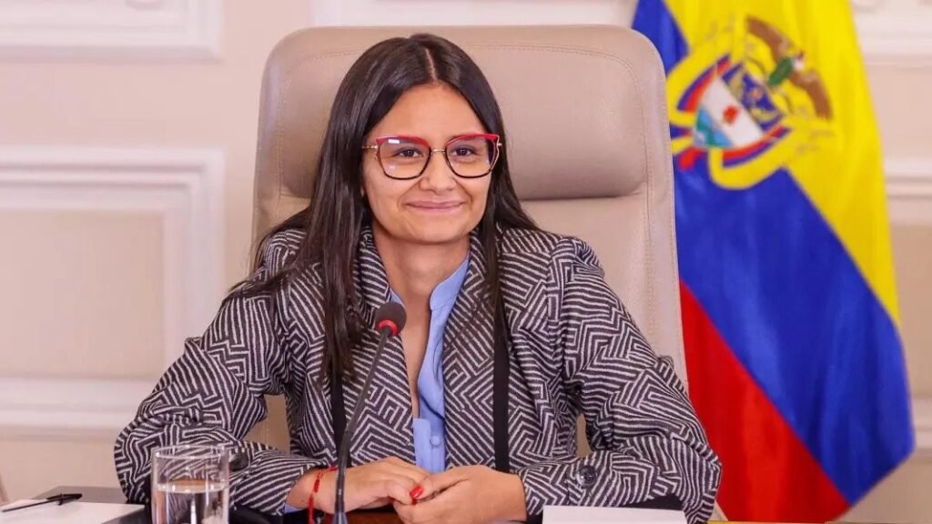 Angie Rodríguez, con gafas y chaqueta de rayas, sonríe sentada ante un micrófono junto a la bandera de Colombia