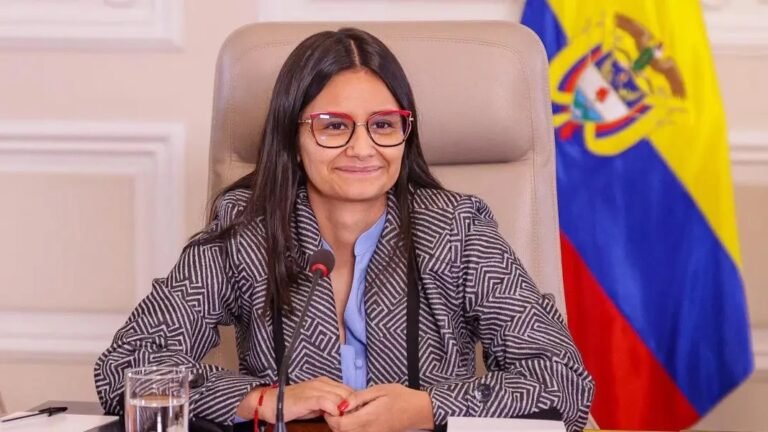 Angie Rodríguez, con gafas y chaqueta de rayas, sonríe sentada ante un micrófono junto a la bandera de Colombia