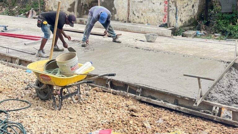 Dos obreros alisan concreto fresco en una calle en construcción, junto a una carretilla amarilla con baldes