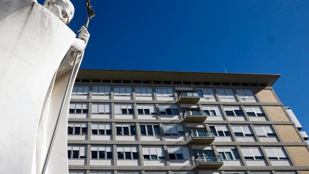 Fachada del Policlínico Gemelli de Roma bajo un cielo azul, con la estatua blanca de un papa en primer plano