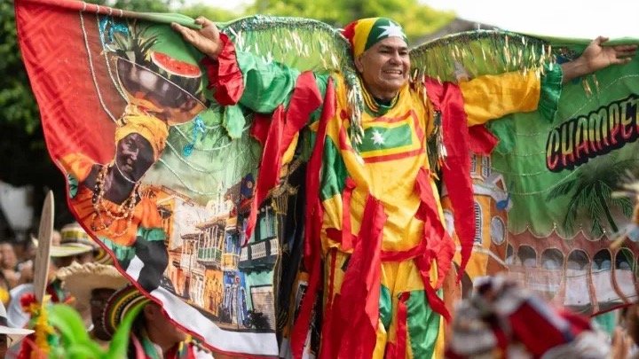 Un hombre con traje de Congo en rojo, amarillo y verde abre los brazos sobre un estandarte de colores durante el desfile del preludio en El Pozón